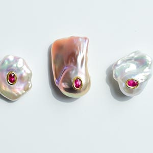 May include: Three iridescent baroque pearls with a gold setting and a vibrant red gemstone. The pearls have a unique, irregular shape and are displayed on a white background. Jewellery.