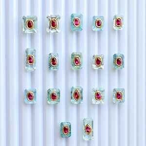 May include: A collection of rectangular aquamarine gemstones, each featuring a central oval ruby and gold setting. The gems are arranged on a white, vertically-lined surface. The gems are of various sizes.