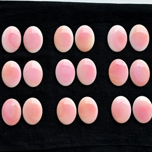 May include: A collection of oval-shaped, polished gemstones in varying shades of pink and peach. The gemstones are arranged in rows on a black surface, showcasing their smooth, reflective surfaces. The overall presentation suggests a focus on jewelry or craft supplies.