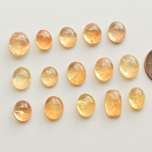 May include: A collection of 15 oval-shaped, translucent, yellow-orange gemstones. The stones are arranged in a random pattern on a white surface. A US penny is included for scale.