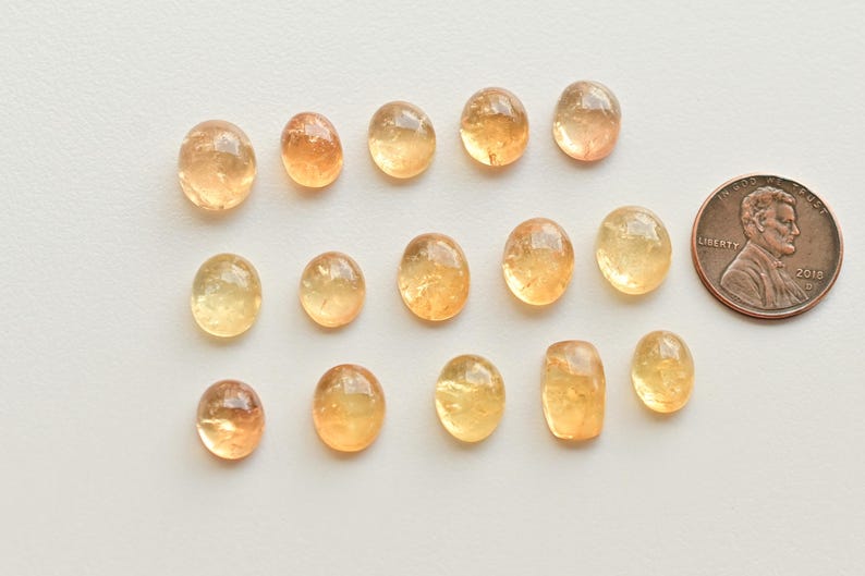 May include: A collection of 15 oval-shaped, translucent, yellow-orange gemstones. The stones are arranged in a random pattern on a white surface. A US penny is included for scale.