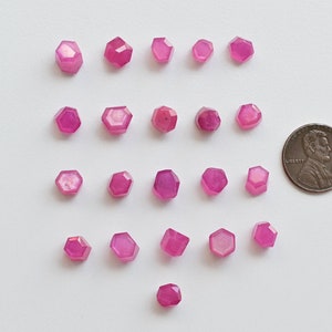 May include: Twenty-four pink faceted gemstones arranged in rows on a white surface. A US penny is included for scale.
