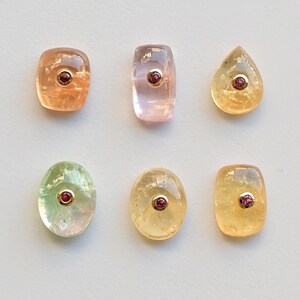 May include: Six different gemstone cabochons, each with a single ruby gemstone set in it. The cabochons are in shades of green, pink, and yellow.