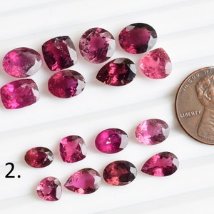 May include: A collection of 15 pink and purple gemstones, some oval, some pear-shaped, and some square, arranged in two rows on a white surface. A penny is included for scale.