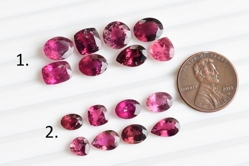 May include: A collection of 15 pink and purple gemstones, some oval, some pear-shaped, and some square, arranged in two rows on a white surface. A penny is included for scale.