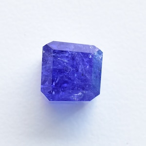 May include: A deep blue, faceted gemstone with a square shape. The gemstone has a slightly cloudy appearance.