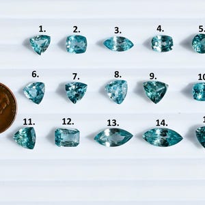 May include: An assortment of fifteen aquamarine gemstones in various shapes, including triangular, square, and oval cuts. A penny is included for size comparison. The gemstones are a light blue color and are arranged on a white surface.