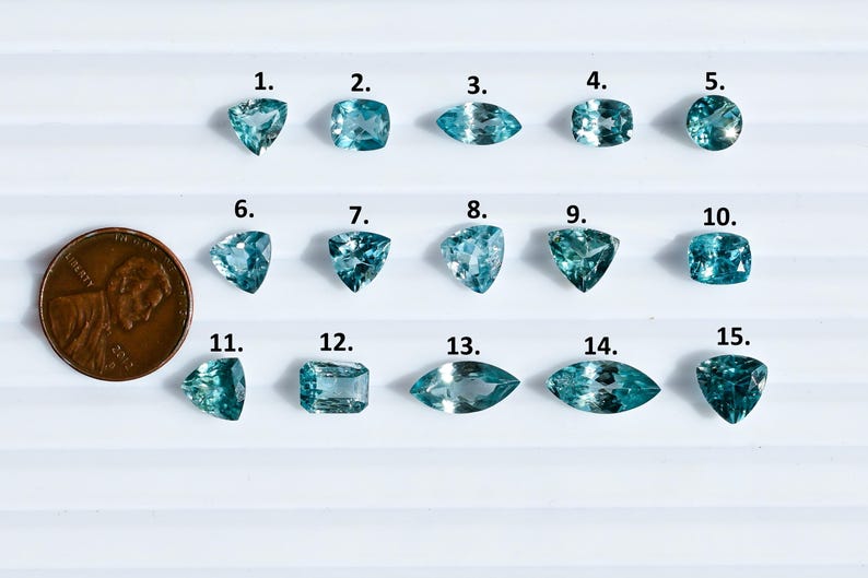 May include: An assortment of fifteen aquamarine gemstones in various shapes, including triangular, square, and oval cuts. A penny is included for size comparison. The gemstones are a light blue color and are arranged on a white surface.