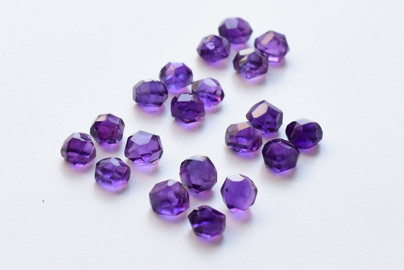 Peut inclure: Un ensemble de perles d'am&eacute;thyste violet fonc&eacute;, facett&eacute;es, dispers&eacute;es sur une surface blanche. Les perles ont une forme g&eacute;om&eacute;trique &agrave; multiples facettes, refl&eacute;tant la lumi&egrave;re. Id&eacute;al pour la cr&eacute;ation de bijoux ou l'artisanat.