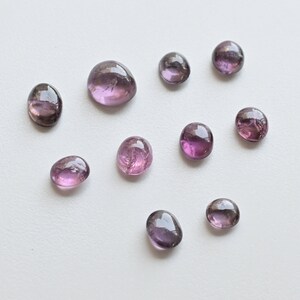 May include: A collection of ten round and oval-shaped, translucent purple gemstones. The stones are arranged in a scattered pattern on a white background.