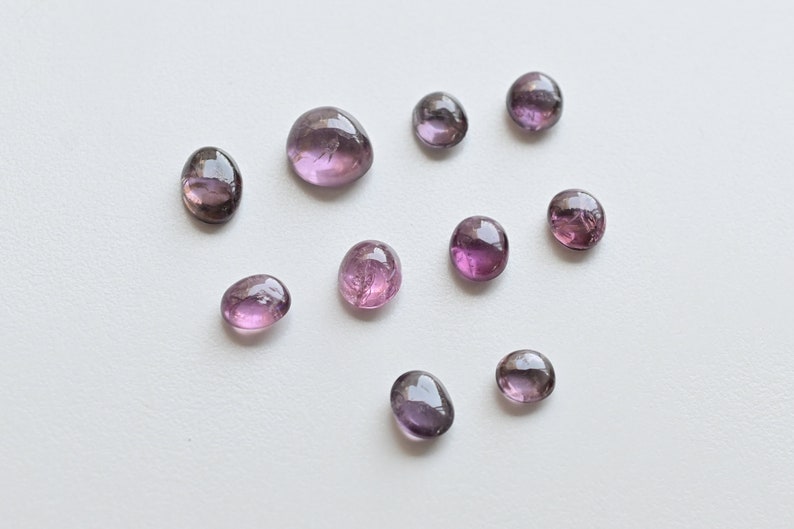 May include: A collection of ten round and oval-shaped, translucent purple gemstones. The stones are arranged in a scattered pattern on a white background.