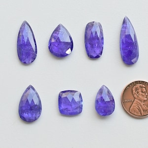 May include: Eight faceted, pear-shaped, purple gemstones, with a US penny for scale.