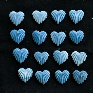 May include: Sixteen pale blue heart-shaped gemstone beads with a textured, fan-like design. The beads are arranged in four rows of four on a black background. The gemstones have a subtle, pearlescent sheen.