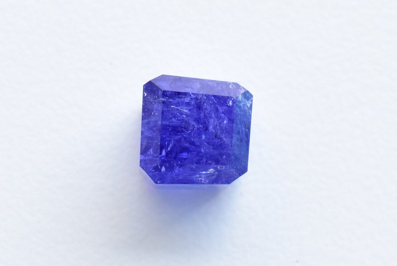 May include: A deep blue, faceted gemstone with a square shape. The gemstone has a slightly cloudy appearance.