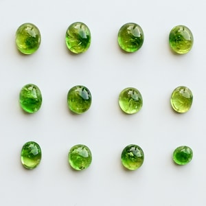 May include: Eleven oval-shaped, translucent green peridot gemstones arranged in a grid pattern. Each stone features a smooth, polished cabochon surface with natural internal inclusions and varying shades of vibrant lime and forest green.