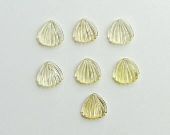May include: Seven translucent, fan-shaped gemstones with a ribbed texture are arranged on a white surface. The gems have a pale yellow hue, and their shape resembles a seashell. The arrangement is symmetrical.