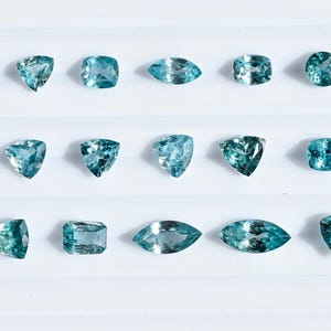 May include: An assortment of aquamarine gemstones in various shapes, including triangular, square, oval, and round cuts. The gems display a range of blue-green hues and are arranged on a white surface. These gemstones are ideal for jewelry making.