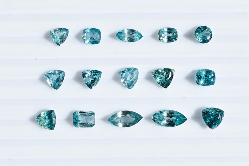May include: An assortment of aquamarine gemstones in various shapes, including triangular, square, oval, and round cuts. The gems display a range of blue-green hues and are arranged on a white surface. These gemstones are ideal for jewelry making.
