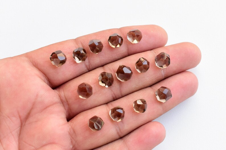 K&ouml;nnte beinhalten: Eine Sammlung facettierter, brauner Perlen. Die Perlen haben eine geometrische Form mit mehreren flachen Oberfl&auml;chen. Die Perlen werden in einer Hand vor wei&szlig;em Hintergrund gehalten. Diese Perlen eignen sich f&uuml;r die Schmuckherstellung.