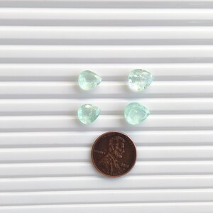 Puede incluir: Cuatro piedras preciosas de color verde claro, facetadas y con forma de pera, dispuestas en formaci&oacute;n cuadrada sobre una moneda de un centavo de d&oacute;lar de cobre, que sirve como referencia de tama&ntilde;o para las peque&ntilde;as piedras transl&uacute;cidas.