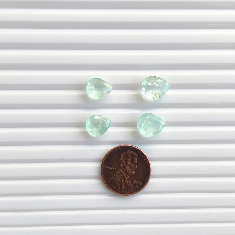 Puede incluir: Cuatro piedras preciosas de color verde claro, facetadas y con forma de pera, dispuestas en formaci&oacute;n cuadrada sobre una moneda de un centavo de d&oacute;lar de cobre, que sirve como referencia de tama&ntilde;o para las peque&ntilde;as piedras transl&uacute;cidas.