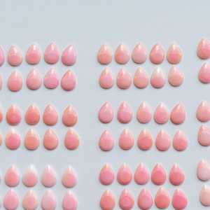May include: Rows of pink teardrop-shaped gemstones arranged on a white surface. The gemstones vary in shades of pink, from light to darker hues. The arrangement suggests a collection of loose stones, possibly for jewelry making or crafting.