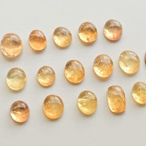 May include: A collection of 15 oval-shaped, translucent, yellow-orange gemstones. The stones are various sizes and have a slightly cloudy appearance.