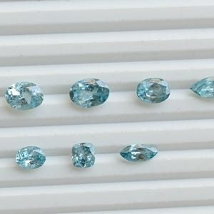 May include: A collection of eight light blue gemstones in various shapes, including oval, round, and pear-cut. The gems are displayed on a white, ridged surface, showcasing their facets and clarity. The gemstones are likely for jewelry making.