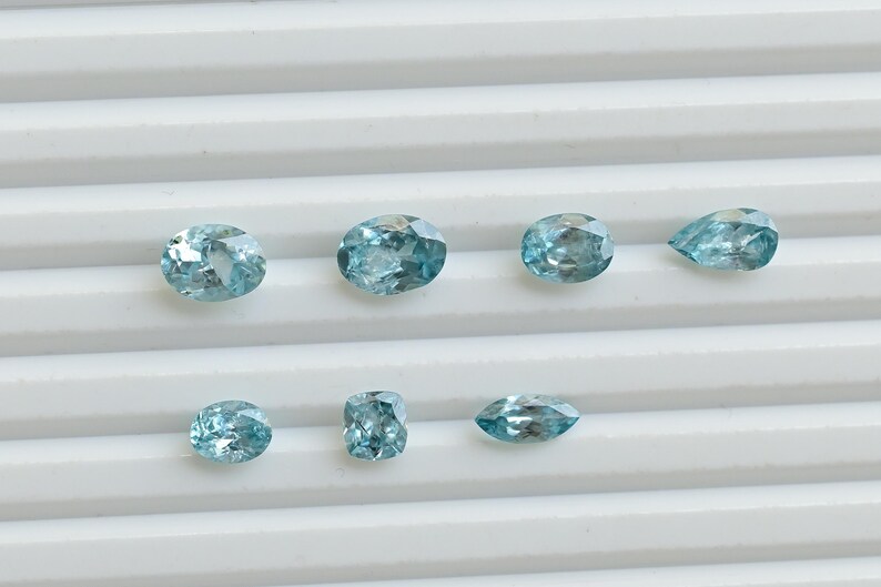 May include: A collection of eight light blue gemstones in various shapes, including oval, round, and pear-cut. The gems are displayed on a white, ridged surface, showcasing their facets and clarity. The gemstones are likely for jewelry making.