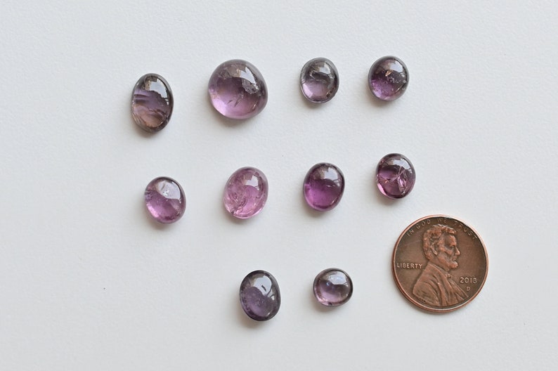 May include: Ten purple and pink oval-shaped gemstones of various sizes, with a penny for scale.