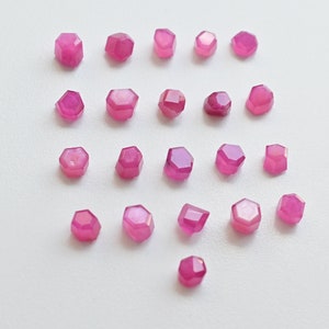 May include: Twenty-five pink faceted gemstone beads, each with a hexagonal shape, arranged in rows on a white surface.