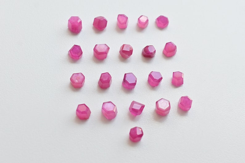 May include: Twenty-five pink faceted gemstone beads, each with a hexagonal shape, arranged in rows on a white surface.