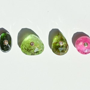 Peut inclure: Quatre pierres précieuses facettées dans des tons de vert et de rose, chacune avec un petit accent central circulaire de couleur or. Les pierres précieuses sont de forme ovale, en goutte et ronde. Les pierres précieuses sont exposées sur une surface blanche.