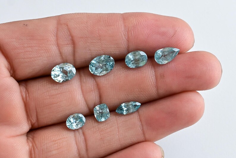 May include: Eight light blue gemstones of different shapes, including oval, pear, and rectangular cuts. The gems are displayed in a hand against a white backdrop, highlighting their clarity and colour.
