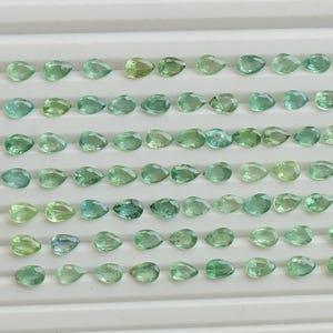 May include: A display of teardrop-shaped gemstones in shades of green, from pale to deep teal. The gems are arranged on a white, ridged surface, highlighting their polished facets and natural colour variations.