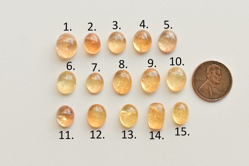 May include: Fifteen yellow-orange oval-shaped gemstones, numbered 1 through 15, are arranged in three rows. A U.S. penny is included for size comparison.