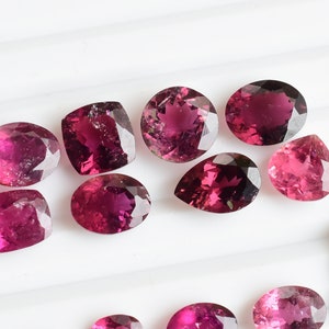 May include: A collection of ten pink and purple gemstones of various shapes and sizes, including oval, pear, and cushion cuts. The gemstones are arranged on a white surface with a penny for scale.