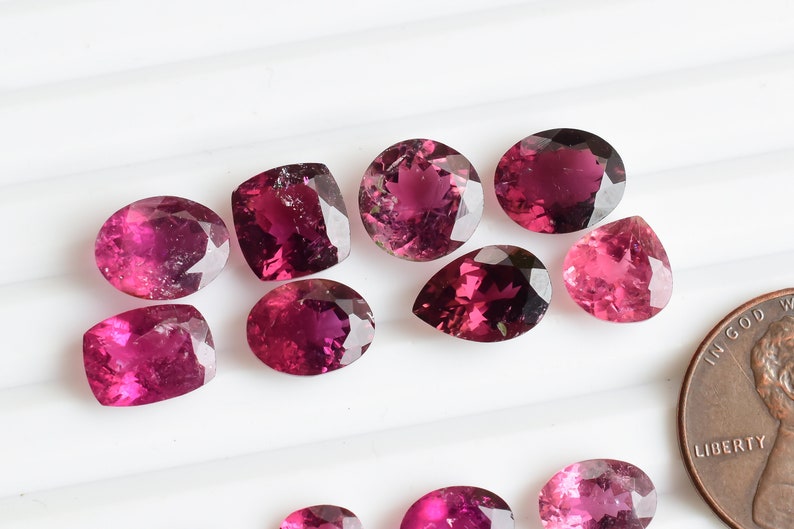 May include: A collection of ten pink and purple gemstones of various shapes and sizes, including oval, pear, and cushion cuts. The gemstones are arranged on a white surface with a penny for scale.