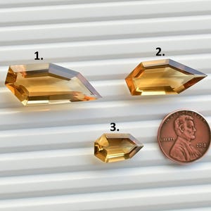 May include: Three faceted, golden-yellow gemstones with sharp angles are displayed. A penny is included for size comparison. The gemstones have a transparent quality, and the light catches their facets. The gems are set against a white, ridged surface.