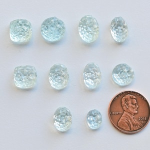 May include: Ten faceted aquamarine gemstones of various sizes and shapes, ranging from oval to square, arranged on a white surface. A UK penny is included for scale.