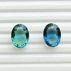 Könnte beinhalten: Zwei oval geschliffene Edelsteine mit einem Farbverlauf von Türkis zu Blau. Die facettierten Edelsteine reflektieren das Licht und sind auf einer weißen, geriffelten Oberfläche platziert. Geeignet für Schmuckherstellung.