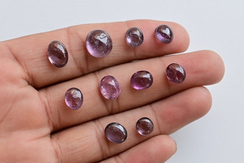 May include: A collection of nine oval-shaped, translucent purple gemstones. The stones are various sizes and have a slight shimmer.