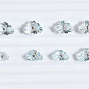 Puede incluir: Ocho gemas en forma de nube, de color azul claro, con un acento central dorado. Las gemas están dispuestas en dos filas de cuatro sobre una superficie blanca. Estas gemas en forma de nube son probablemente para joyería.