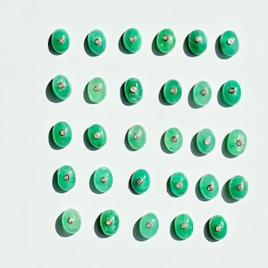 Könnte beinhalten: Eine Anordnung ovaler, grüner Edelsteine, jeweils mit einem kleinen, silberfarbenen Kreis in der Mitte. Die Edelsteine sind in einem Raster auf weißem Hintergrund angeordnet und erzeugen ein ansprechendes Muster.