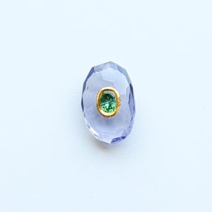 May include: A light purple, oval-cut gemstone with a gold setting. The centre features a smaller, green gemstone. The gem is set against a white background.