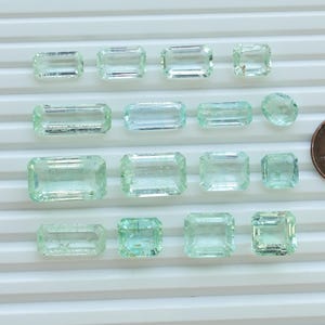 May include: An assortment of aquamarine gemstones in various rectangular and oval shapes, displayed on a white surface. A US penny is included for size comparison. The gemstones exhibit a pale blue-green hue and are faceted.