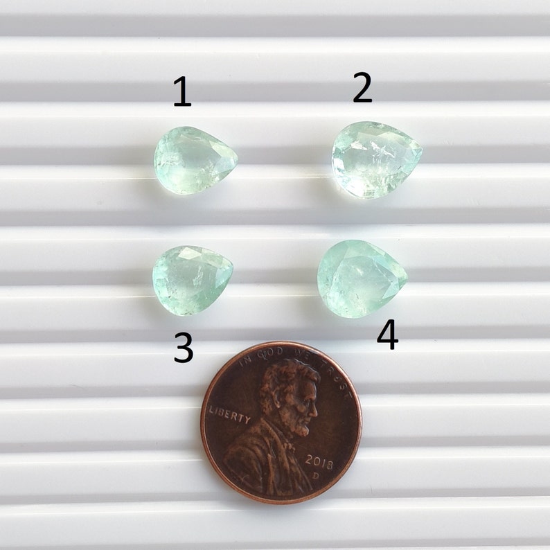 Puede incluir: Cuatro piedras preciosas verdes en forma de pera, numeradas del 1 al 4, se muestran sobre una superficie blanca con un centavo para la escala.