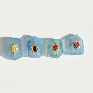May include: Four light blue gemstone pendants with gold settings. Two pendants have a gold centre, and two have a red centre. The pendants are arranged in a row on a white surface.
