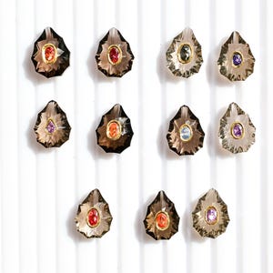May include: An array of eleven gemstone pendants with various colored center stones, including red, orange, purple, and blue. Each pendant has a gold-colored bezel and a smoky quartz-like outer stone. The pendants are arranged on a white, vertically-lined background.