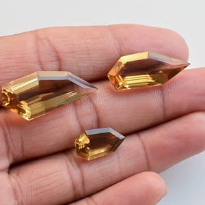 May include: Three faceted, golden-yellow gemstones with pointed ends are displayed in a hand. The gems have a rectangular shape with angled sides, catching the light to reveal their internal structure. The gems vary in size.